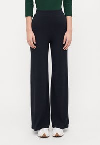 Pantalons larges noirs avec des rayures verticales, texture douce et taille haute, associés à des chaussures de sport blanches.