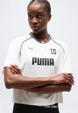 Naine, kellel on patsidega juuksed ja valge Puma sportlik särk numbriga 10, käed rüpes, vaatab küljele ühevärvilise tausta ees.