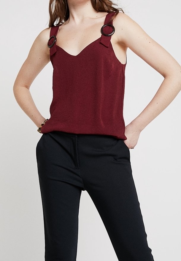 Femme portant un top sans manches rouge foncé avec des détails en anneaux sur les bretelles et un pantalon noir, mains sur les hanches devant un fond uni.