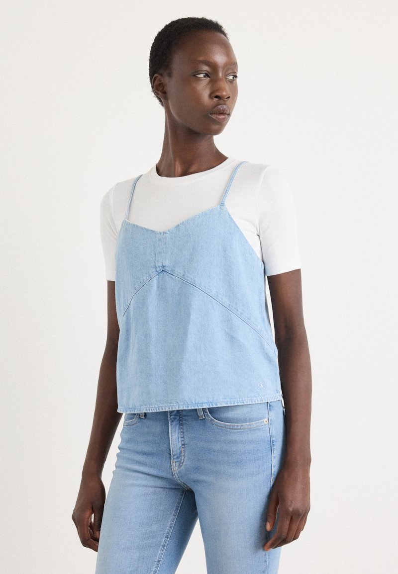 Light blue denim camisole with thin straps, a V-neckline, and seam detailing, paired with a white short-sleeve top and light blue jeans.