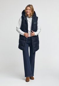 Marinblå puffväst med huva, med dragkedja fram, sidofickor och en slät textur. Kombineras med en ljusgrå sweatshirt och högmidjade jeans.