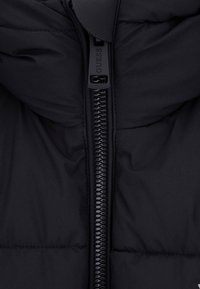 Juodas pūkinis švarkas su aukštu apykakle, turintis visiškai užtrauktuką ir siuvinėtą tekstūrą. Lygus, matinio paviršiaus ir minimalus detalizavimas.