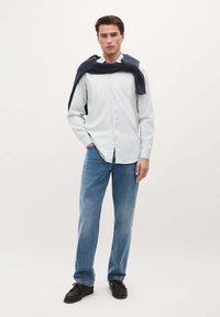 Cămașă bleu deschis cu nasturi, blugi blue denim, pantofi negri și un pulover bleumarin pus pe umeri. Modelul stă pe un fundal neutru.
