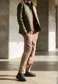 Chaqueta verde acolchada, camisa de botones color óxido, pantalones beige con textura gruesa y zapatillas negras. Materiales texturizados y un look en capas.