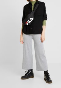 Schwarze knopffreie Hemd, grüner Rollkragenpullover, graue gerippte weite Hosen, schwarze Schnürstiefel und schwarze Fila Umhängetasche mit weißem Logo.