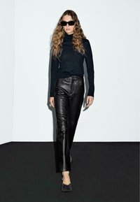 Donna che cammina sulla passerella indossando un dolcevita nero, pantaloni di pelle nera, tacchi neri e occhiali da sole neri di grandi dimensioni, contro un muro bianco e un pavimento nero.