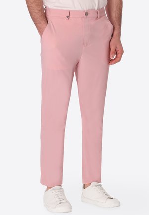 Uomo con pantaloni slim rosa chiaro e sneakers bianche, in piedi con le mani nelle tasche davanti a uno sfondo semplice.