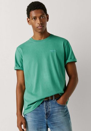 Ung mand iført en grøn T-shirt med rullede ærmer og blå jeans, med den ene hånd i lommen, stående mod en enkel, lys baggrund.