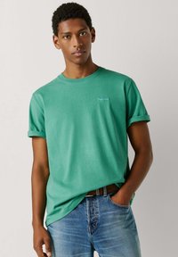 Noor mees, kes kannab rohelist varrukaid üles keeratud t-särki ja siniseid teksaseid, ühe käega taskus, seisab heleda ühevärvilise tausta ees.