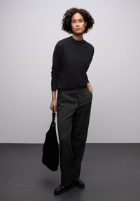 Černý svetr s vysokým límcem, kombinovaný s texturovanými tmavými kalhotami. Modelka drží černou tašku, stojí na šedé podlaze s jednobarevným pozadím.