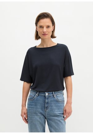 Vrouw met kort bruin haar staat frontaal, draagt een losse zwarte T-shirt en een blauwe spijkerbroek tegen een effen witte achtergrond.