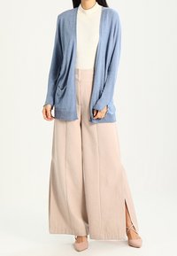 Kvinna klädd i ljusblå cardigans, vit ribbad topp, vida beige byxor med sidofickor, och beige spetsiga skor med ankelsnören.