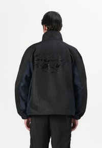 Black and navy jacket featuring a high collar, elastic cuffs, and a glossy graphic on the back, with a smooth, lightweight texture.
