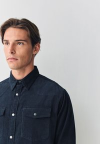 Mørkeblå skjorte med knapper lavet af tekstureret stof, med to brystlommer med trykknapper og en spids krave.