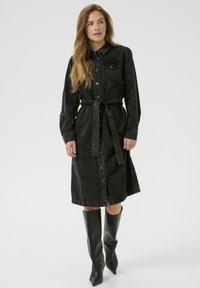Robe chemise en denim noir avec des manches longues, boutonnée à l'avant et ceinturée à la taille. Elle est dotée d'une poche poitrine et d'un ourlet au niveau des genoux. Texture lisse.