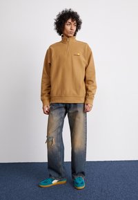 Pullover de lã castanho com gola alta e fecho éclair à frente, combinado com jeans claros desgastados e ténis azul-piscina com solas de borracha.