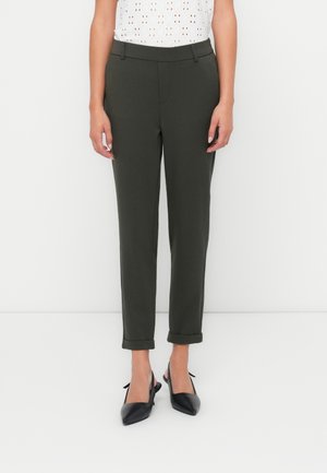 Vrouw die donkergroene enkellange broek met omslag en riemlussen draagt, gecombineerd met zwarte slingback-hakken met puntige neus, staand tegen een witte achtergrond.