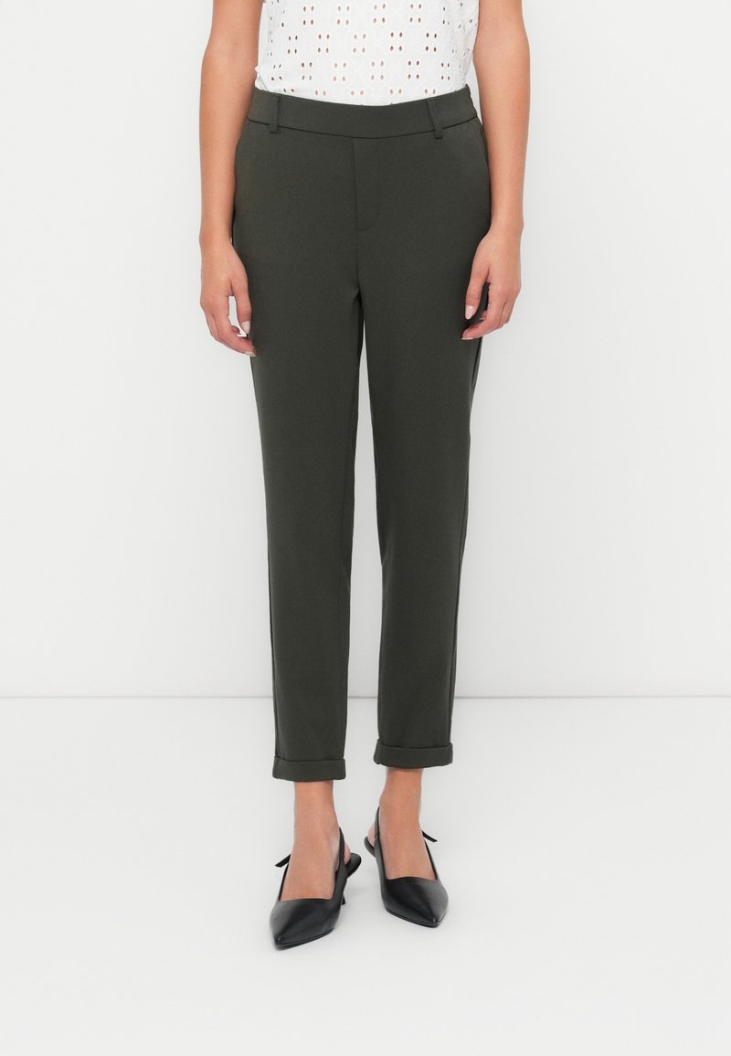 Femme portant un pantalon vert foncé retroussé à la cheville avec passants de ceinture et des escarpins slingbacks noirs à bouts pointus, debout sur un fond blanc.