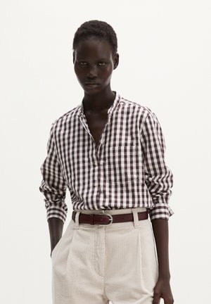 Femme portant une chemise à manches longues à carreaux marron et blanc rentrée dans un pantalon en velours côtelé beige avec une ceinture marron foncé.