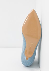 Chaussure à talon haut en denim bleu à bout pointu, semelle lisse couleur fauve et petit talon, photographiée par le dessous sur fond blanc.