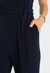 Mörkblå jumpsuit med en slät yta, knytdetalj vid midjan och en sidoficka. Inkluderar ett lätt guldarmband på handleden.