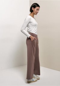 Femme debout de profil, les mains dans les poches d'un pantalon marron à jambes larges, portant un haut blanc à manches longues et des baskets blanches avec des accents bleus.