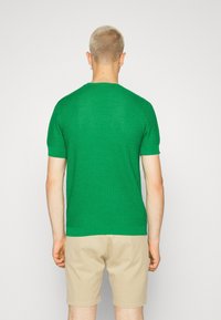 Grön stickad kortärmad tröja med en strukturerad mönster. Nederkanten och ärmsluten är enfärgade gröna. Bärs med beige shorts.