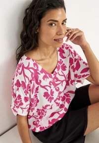 T-shirt met korte mouwen, met een witte basis en een groot roze bloemenpatroon. De halslijn heeft een diepe V-vorm en de stof heeft een textuurafwerking.
