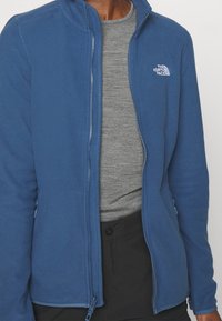 Niebieska kurtka polarowa z zamkiem z przodu i stójką. Pod spodem szara koszula z długim rękawem. Logo na górnej części klatki piersiowej oraz kieszenie na suwak po bokach.