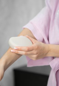 Witte ergonomische handmassager tegen een huidkleurige arm, met een gladde textuur. De persoon draagt een lichtroze badjas, achtergrond vervaagd.