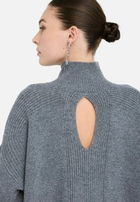 Vrouw met bruin haar in een knot draagt een grijze ribgebreide trui met een sleutelgatdetail aan de achterkant en lange zilveren kettingoorbellen, kijkt naar links.