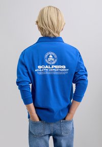 Niebieska koszulka polo z długim rękawem i kołnierzykiem, z białym napisem i grafiką na plecach, zestawiona z jasnoniebieskimi dżinsami.