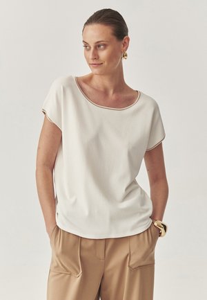 Kvinde iført en hvid kortærmet bluse med beige kant, beige bukser med lommer, guldøreringe og et guldarmbånd, hænderne i lommerne.