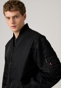 Νεαρός άνδρας με σγουρά μαλλιά που φοράει μαύρο μπουφάν τύπου bomber με τσέπη με φερμουάρ στο αριστερό μανίκι, στέκεται μπροστά σε απλό φόντο.