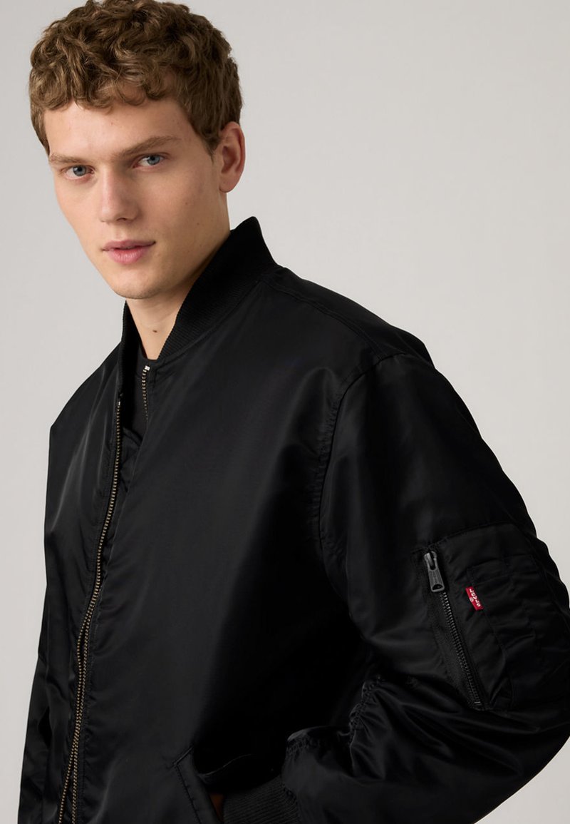 Giovane uomo con capelli ricci che indossa un bomber nero con una tasca con cerniera sulla manica sinistra, in piedi contro uno sfondo neutro.