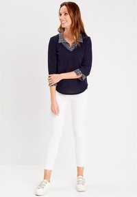 Marineblauer Pullover mit V-Ausschnitt und weißem, blumenmustertem Kragen und Bündchen; kombiniert mit weißen, schmal geschnittenen Hosen und weißen Sneakern.