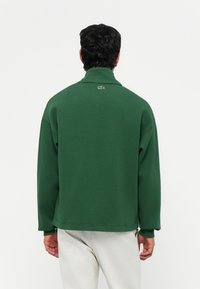 Groene sweatshirt met een hoge kraag, verlaagde schouders en zijritsen. Heeft een gestructureerde stof en een klein logo op de achterkant.