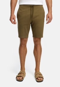 Homme portant un short olive à cordon et des sandales en suède beige, se tenant devant un fond blanc uni.