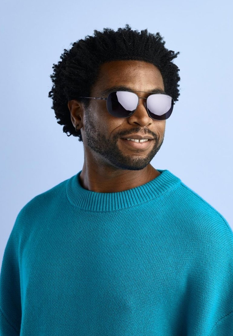 Occhiali da sole neri con lente riflettente, forma esagonale; indossati con un maglione a maglia turchese con un collo rotondo e a coste.