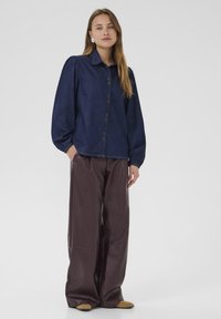 Denim button-up shirt with puffed sleeves, dark blue color, paired with wide-leg brown leather pants and beige slip-on shoes.
