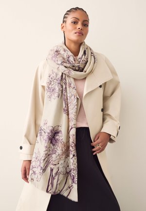 Şal bej cu motive florale și desene cu linii violet, purtat peste o bluză roz pal și o trench coat crem, pe un fundal neutral.