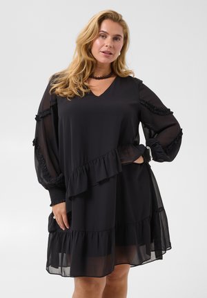 Femme aux longs cheveux blonds portant une robe noire à volants avec des manches bouffantes transparentes et un collier sombre orné de perles sur un fond uni.