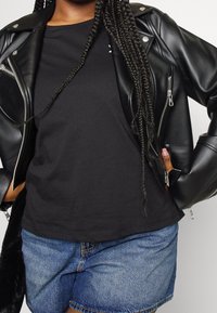 Czarna kurtka motocyklowa z eko skóry z srebrnymi zamkami błyskawicznymi i zatrzaskami, zestawiona z czarną koszulką i dżinsowymi szortami. Widać splecione włosy.