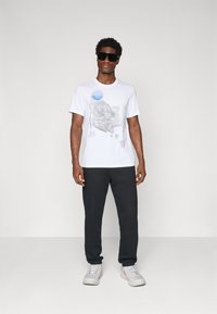 Hvid T-shirt med grafisk design med blå elementer, parret med sorte sweatpants og grå sneakers i en minimalistisch kontekst.