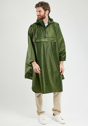 Homme portant un poncho de pluie à capuche vert olive, pantalon beige et baskets blanches, debout devant un fond blanc, regardant sur le côté.