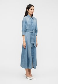 Φόρεμα από τζιν σε ανοιχτό μπλε, με γιακά, μανίκια μέχρι τον αγκώνα, ζώνη στη μέση και μήκος μέχρι τον αστράγαλο με σκίσιμο στο μπροστινό μέρος.