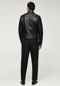 Uomo in piedi, visto da dietro, che indossa una giacca di pelle nera, pantaloni neri e scarpe eleganti nere su uno sfondo chiaro e semplice.