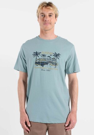 T-shirt en coton bleu clair avec un motif graphique représentant des palmiers, une camionnette et le texte : "Vacances permanentes depuis 1985" en couleurs contrastées.