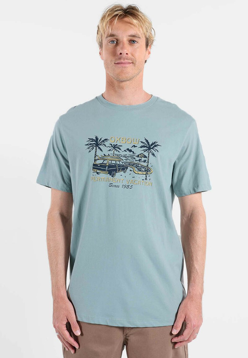 T-shirt en coton bleu clair avec un motif graphique représentant des palmiers, une camionnette et le texte : "Vacances permanentes depuis 1985" en couleurs contrastées.