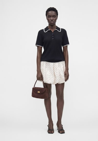Femme portant un polo noir à manches courtes avec un liseré blanc, une jupe blanche à motifs, des sandales noires, et tenant un petit sac à main marron.